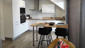 A modern kitchen with white cabinets, a black oven, and a stainless steel range hood. There is a dining area with a wooden table and black chairs. - Guidel. Superb apartment with large terrace. (Guidel)
