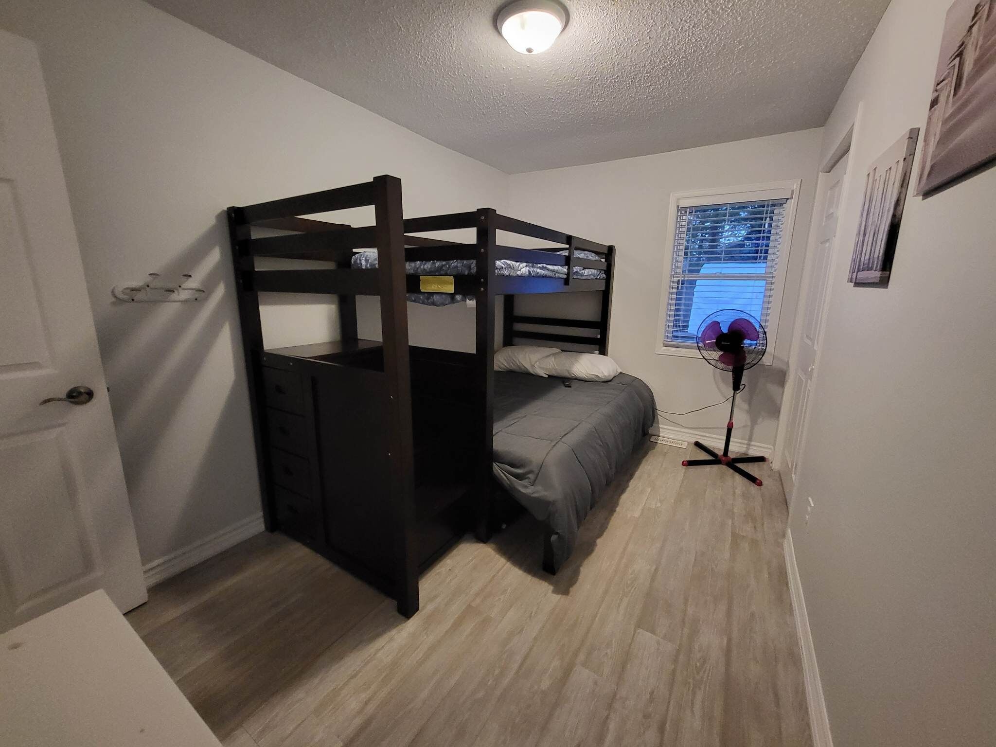 Una habitación pequeña con una litera de madera, una mesita de noche, un pequeño cajón, un ventilador de pie y una ventana con persianas.