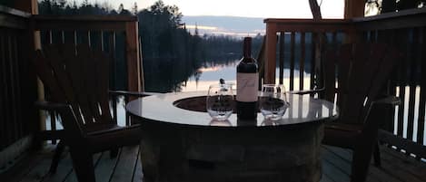 A stone table with a bottle and glasses on a wooden deck overlooking a lake.