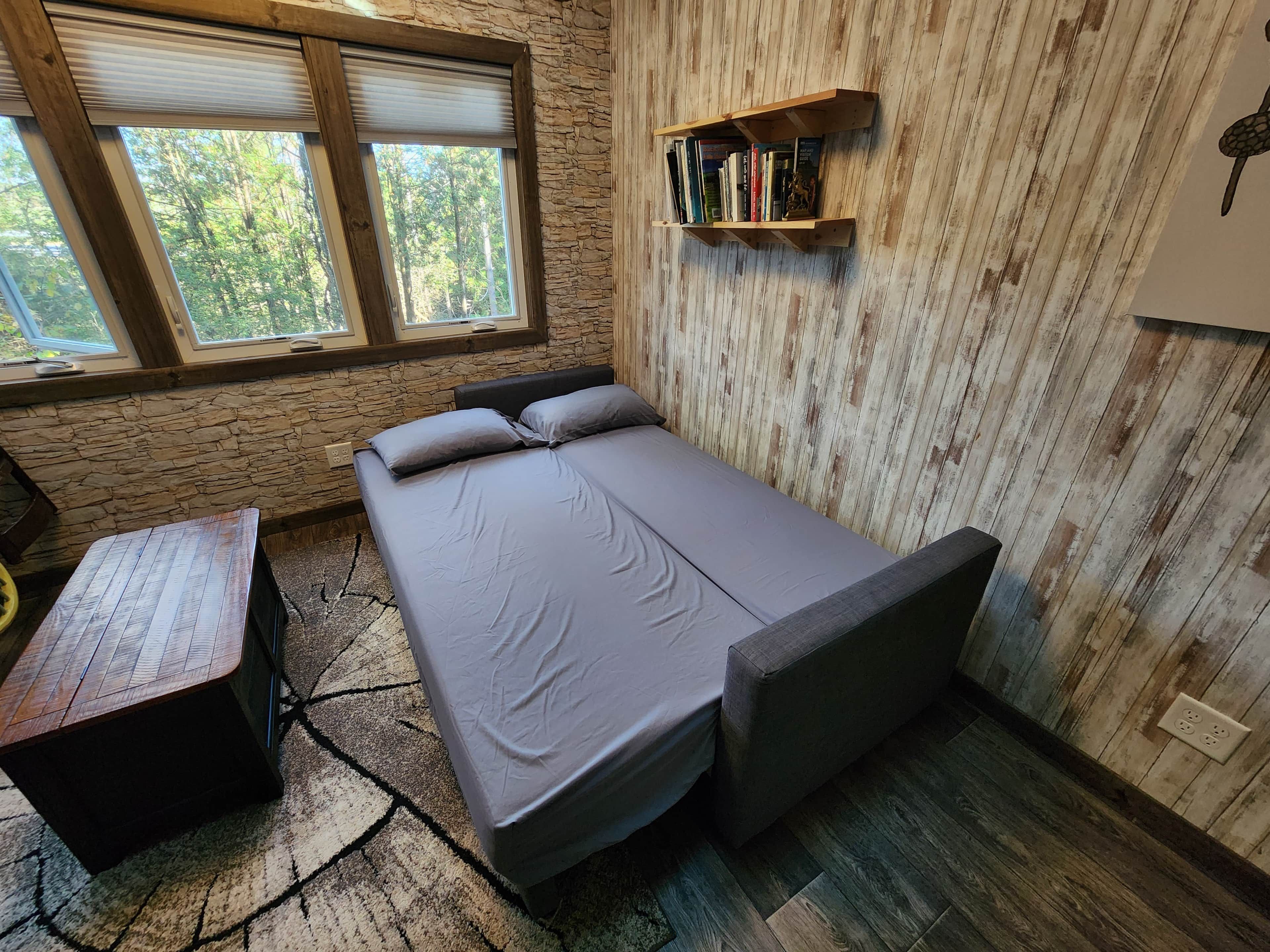 Una stanza compatta con un letto, un tavolino, una libreria e una finestra vista alberi.