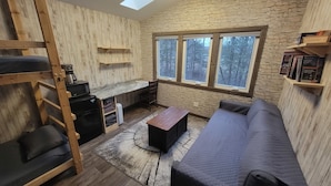 Acogedora habitación tipo cabaña con literas, una pequeña kitchenette, un sofá y un ventanal con vistas al bosque.