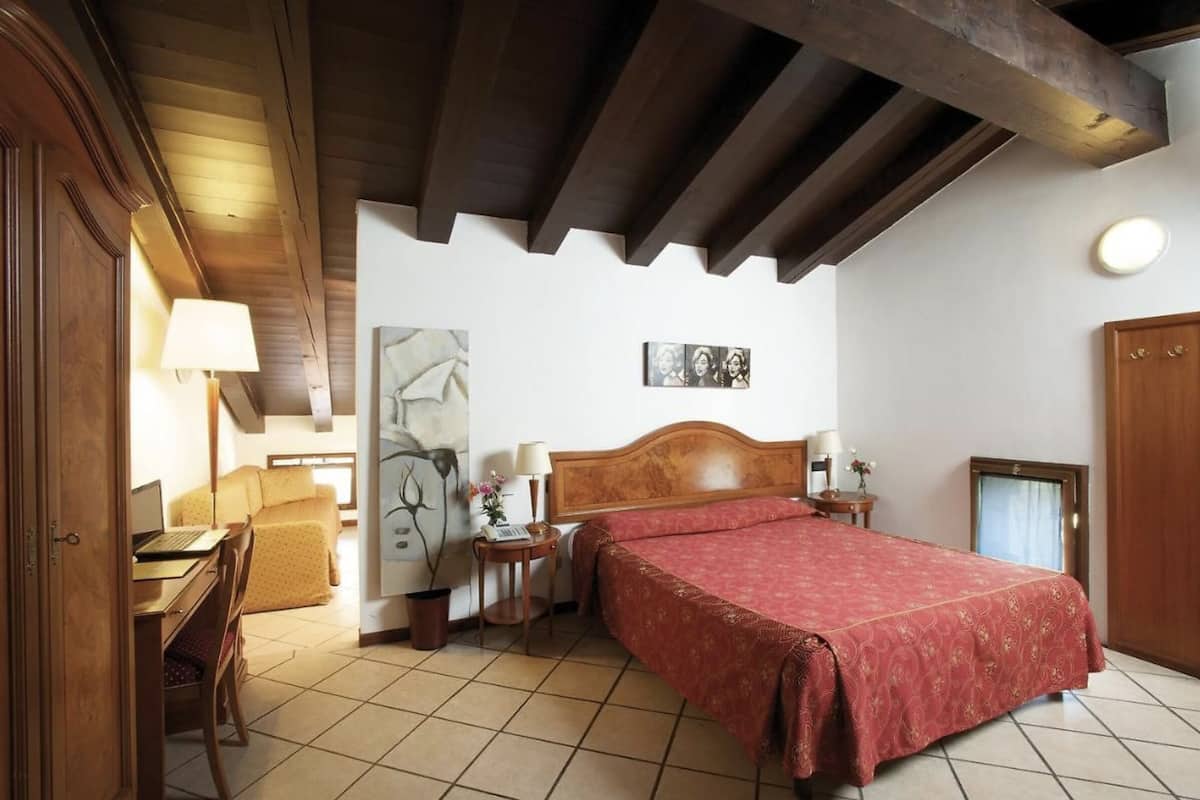 Un dormitorio con una cama de madera, un escritorio con una computadora portátil, una silla, una mesita de noche con una lámpara y una pared decorada con un cuadro enmarcado.