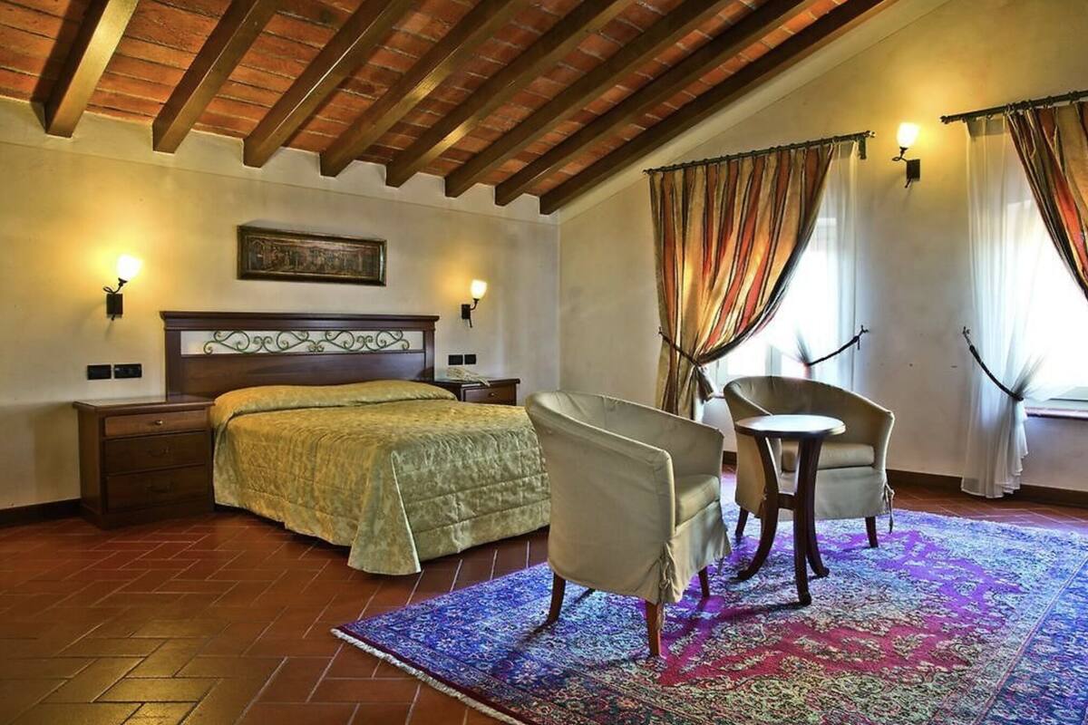 Un dormitorio con techo de madera, una cama con colcha verde, dos sillas, una mesita y una alfombra colorida.