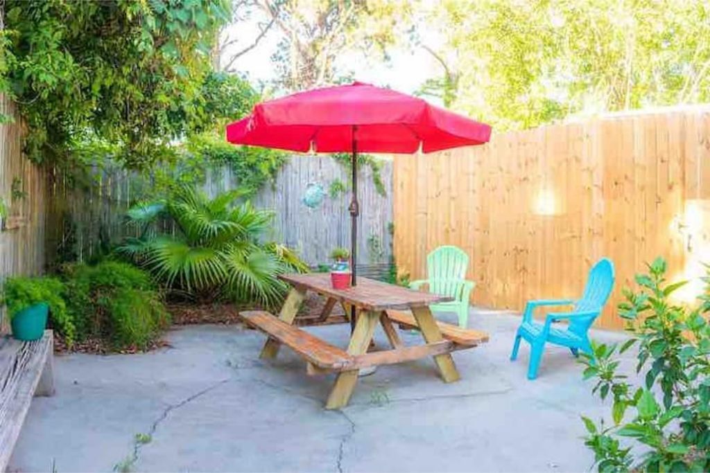 A wooden picnic table with benches and a red umbrella, surrounded by a wooden fence and greenery.