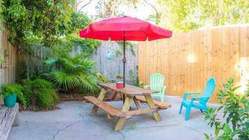 A wooden picnic table with benches and a red umbrella, surrounded by a wooden fence and greenery.
