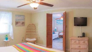 A bedroom with a bed, a ceiling fan, a television, a wicker chair, a dresser, and a door leading to another room.