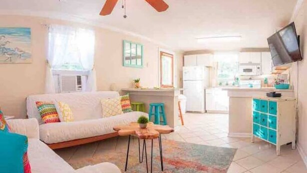 A compact living space with a sofa, a small table, a ceiling fan, a kitchen area, and a TV mounted on the wall.
