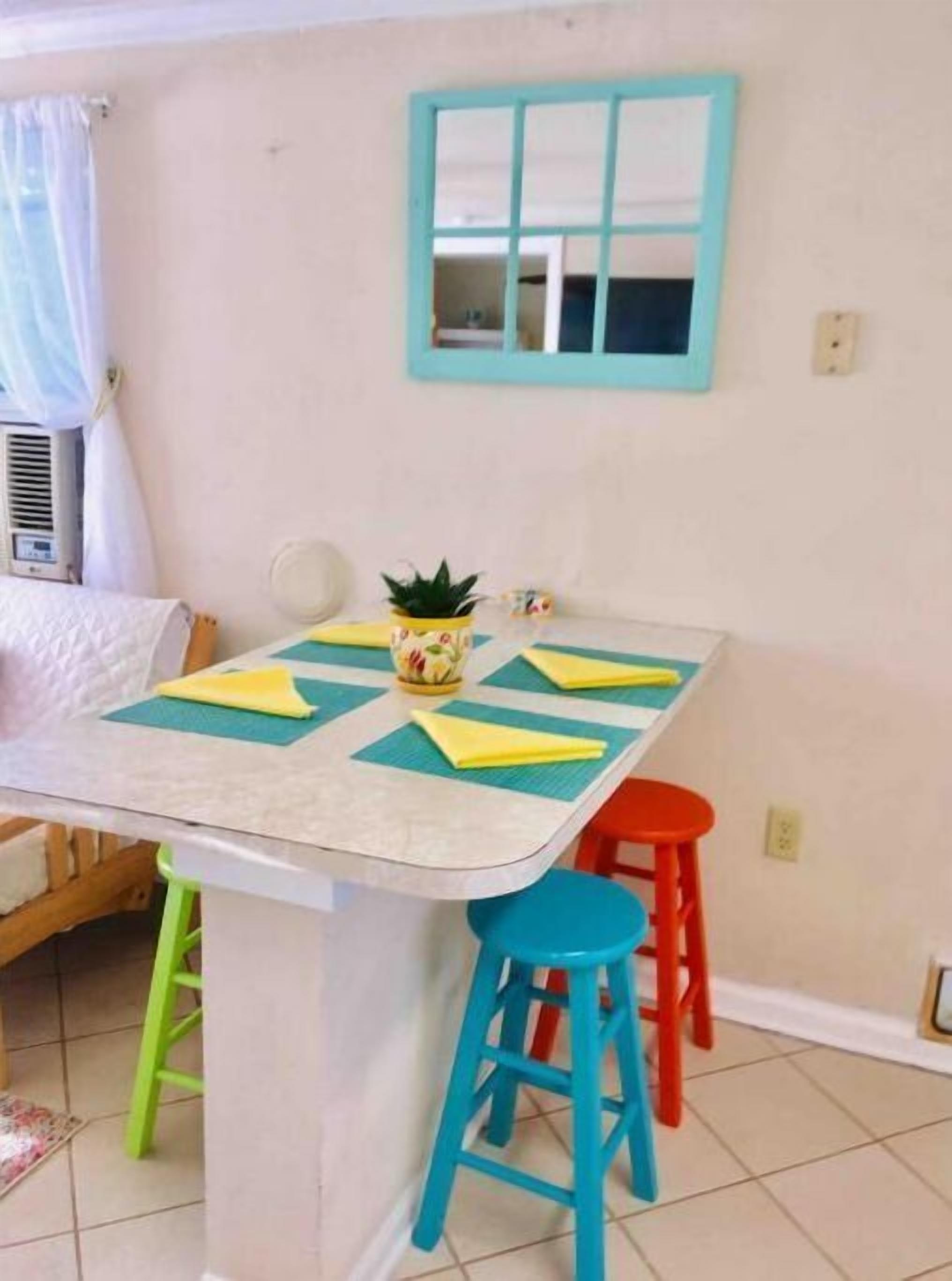 A small dining area with a table, colorful stools, a potted plant, and a window with a blue frame.