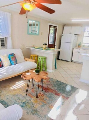 Un espace de vie compact comprenant un canapé, une petite table basse, un ventilateur de plafond et un coin cuisine avec un réfrigérateur et des tabourets.