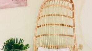 A wicker chair with a white cushion, a small round table with a blue pot of a green plant, and a painting on the wall.