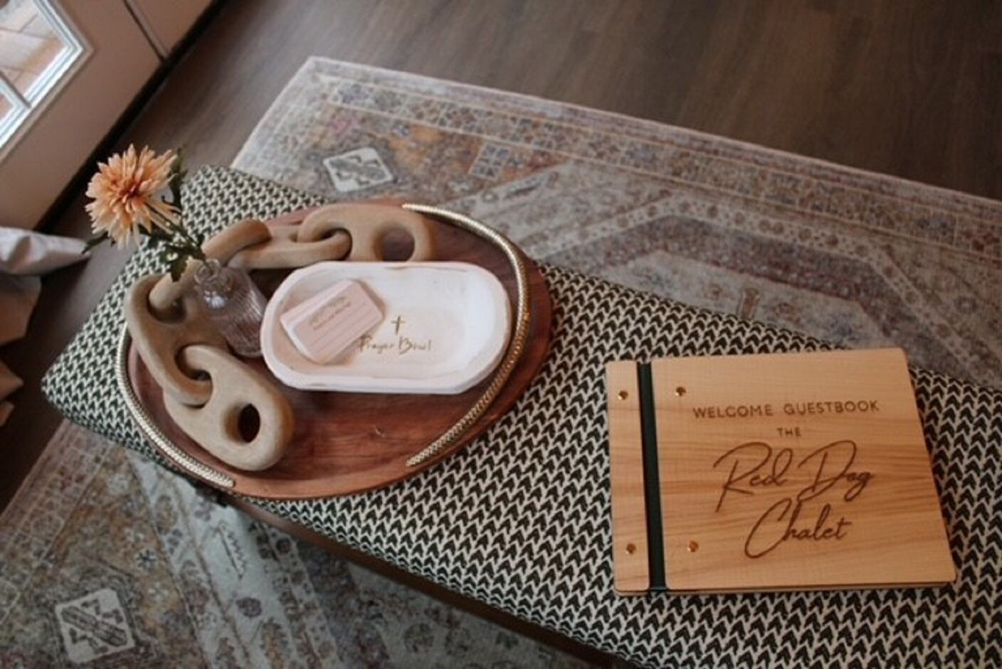 Un plateau en bois avec un porte-savon et une fleur décorative, posé sur un tapis à motifs. Une pancarte en bois indique : « LIVRE D’OR – BIENVENUE AU Red Dog Chalet ».