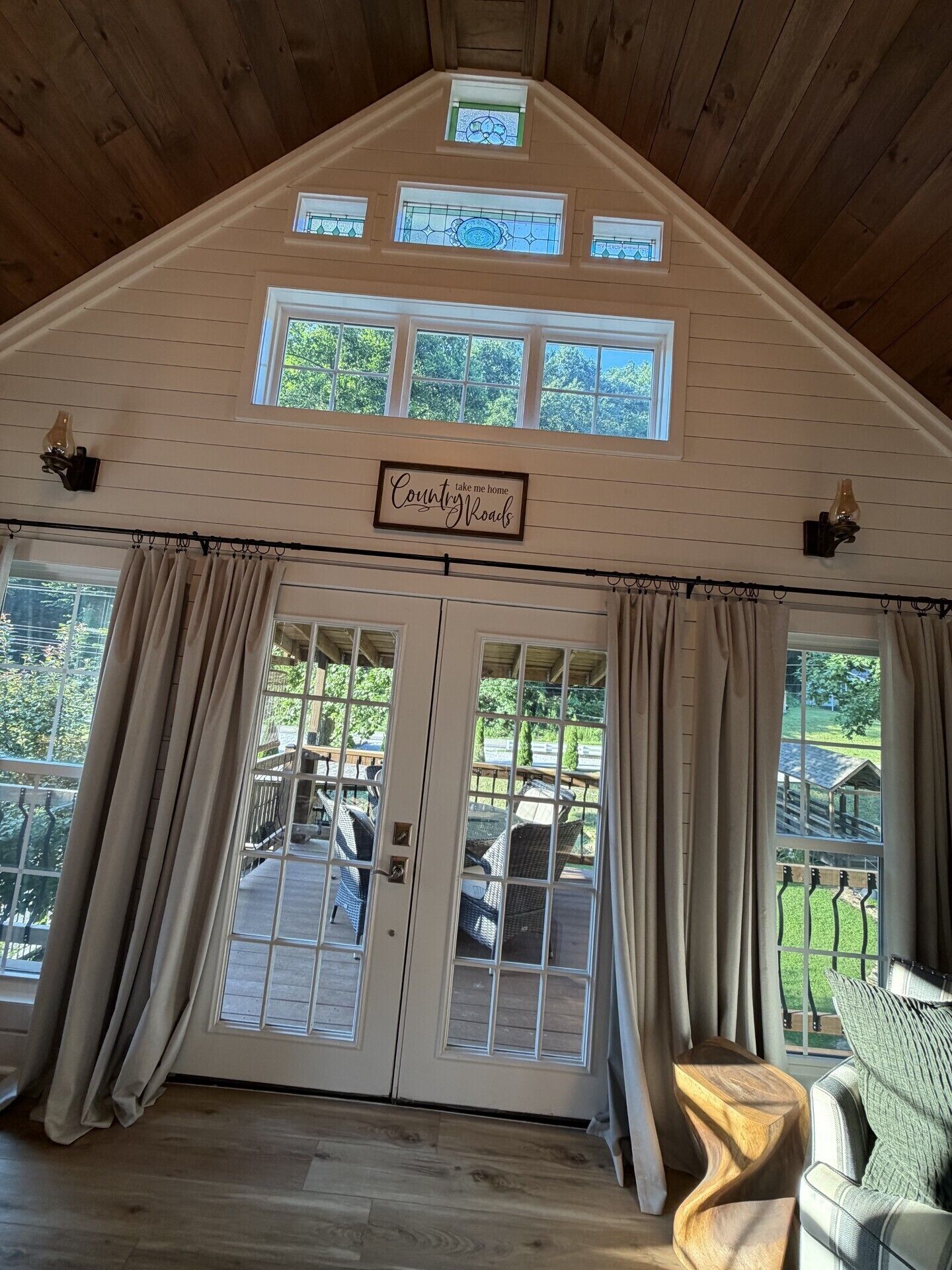 A room with a wooden ceiling, a sign reading 'Country Roads,' large windows with curtains, and a view of outdoor furniture.