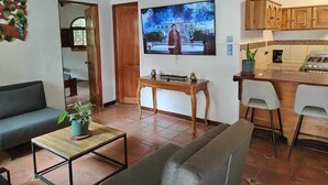 A living room with a sofa, a coffee table, and a TV mounted on the wall. There is a kitchen area with a stove and cabinets. - Hotel Planeta Sano (Quepos)