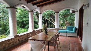 A covered patio with a wooden table and chairs, a sofa, and a view of lush greenery. - Hotel Planeta Sano (Quepos)