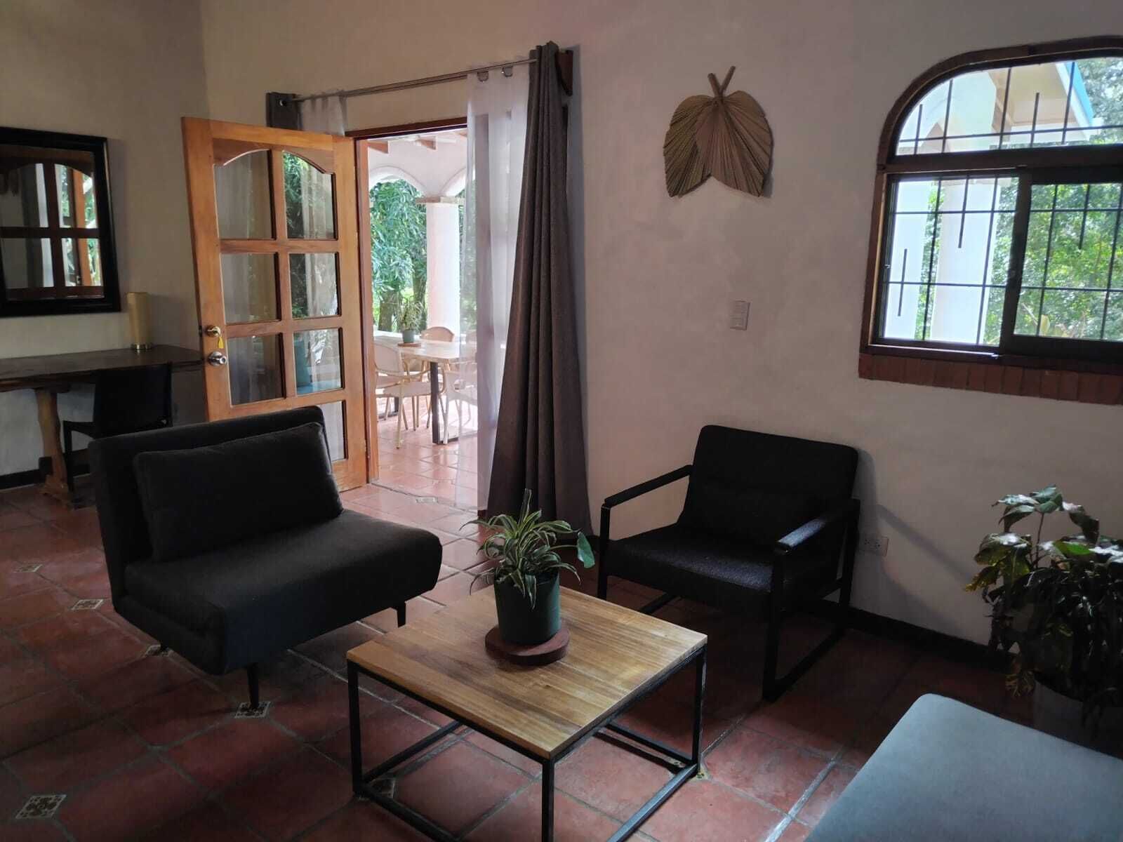 A room with a sofa, a chair, a small table with a plant, a window with bars, and a door leading to an outdoor area.