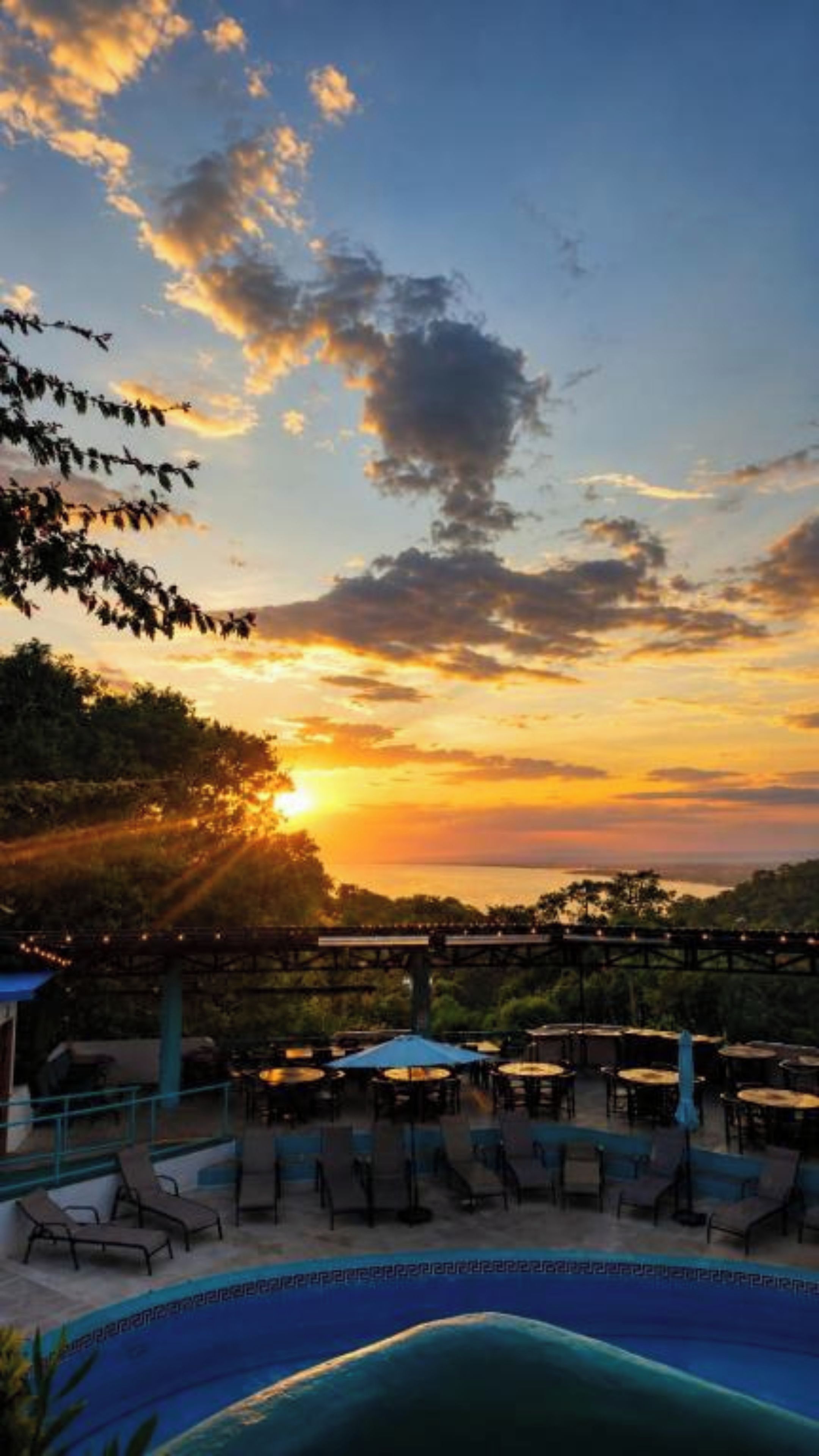 A poolside area with lounge chairs and umbrellas, overlooking a body of water during sunset.