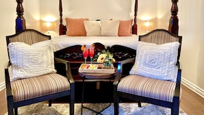 A bedroom with a four-poster bed, two upholstered chairs, a tray with fruit and drinks, and two wall-mounted lamps.