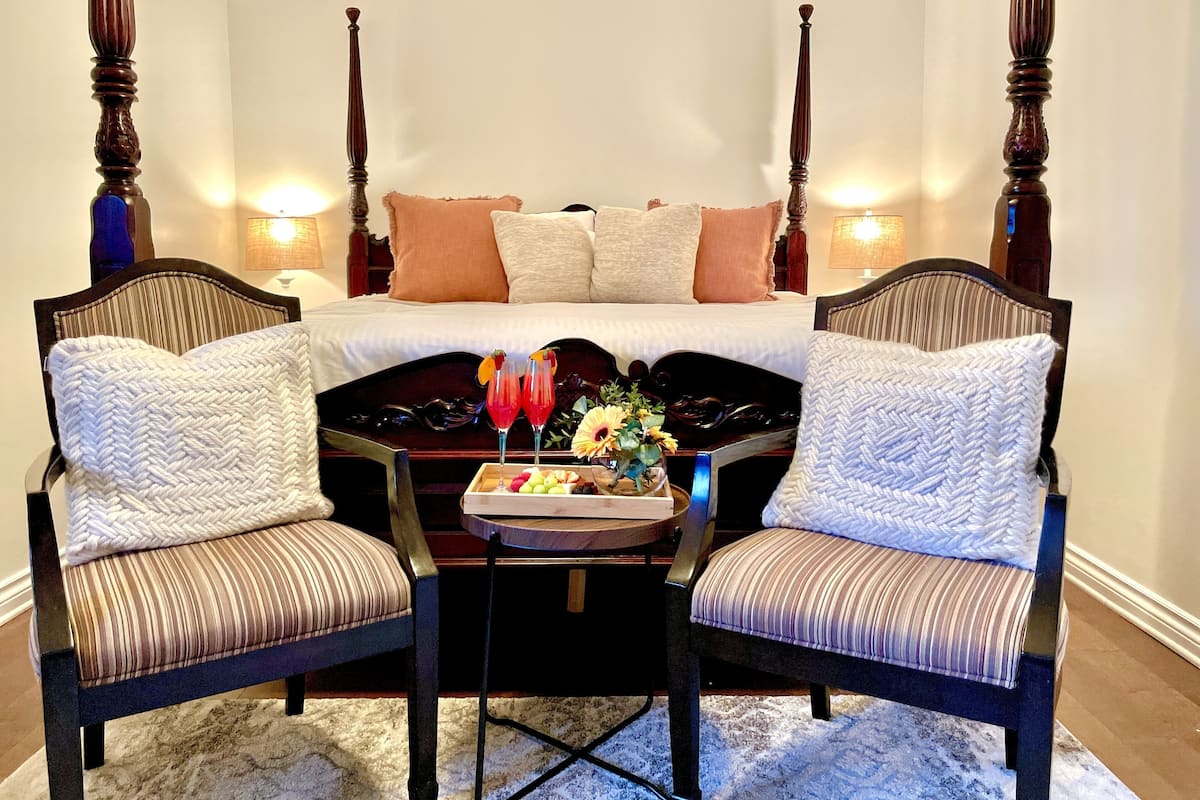 Un dormitorio con cama con dosel, dos sillones tapizados, una bandeja con fruta y bebidas, y dos lámparas fijadas a la pared.