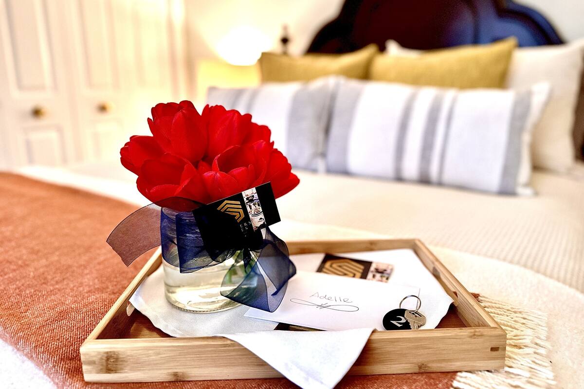 Una bandeja de madera con un jarrón de flores rojas, una tarjeta negra y un llavero sobre una cama con almohadas a rayas.