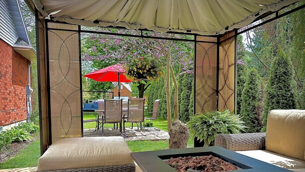 A covered patio area with outdoor furniture, a fire pit, and a view of a backyard with a red umbrella and a tree.