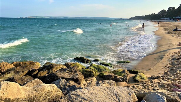 A beach with rocky foreground, clear blue water, and people in the distance. - Gulliver's (Poole)