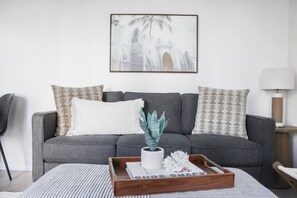 A grey sofa with patterned cushions, a framed picture above, a wooden tray with a plant, and a striped rug.
