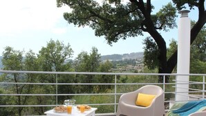 Un balcone con un tavolo apparecchiato per la colazione, una sedia bianca con un cuscino giallo e vista su un paesaggio verde.