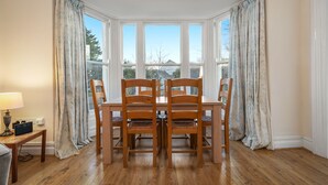 A dining area with a wooden table and chairs, large windows with curtains, and a lamp on a side table. - Apartment 4 St Mary's House - Short Walk to Beach (Tenby)