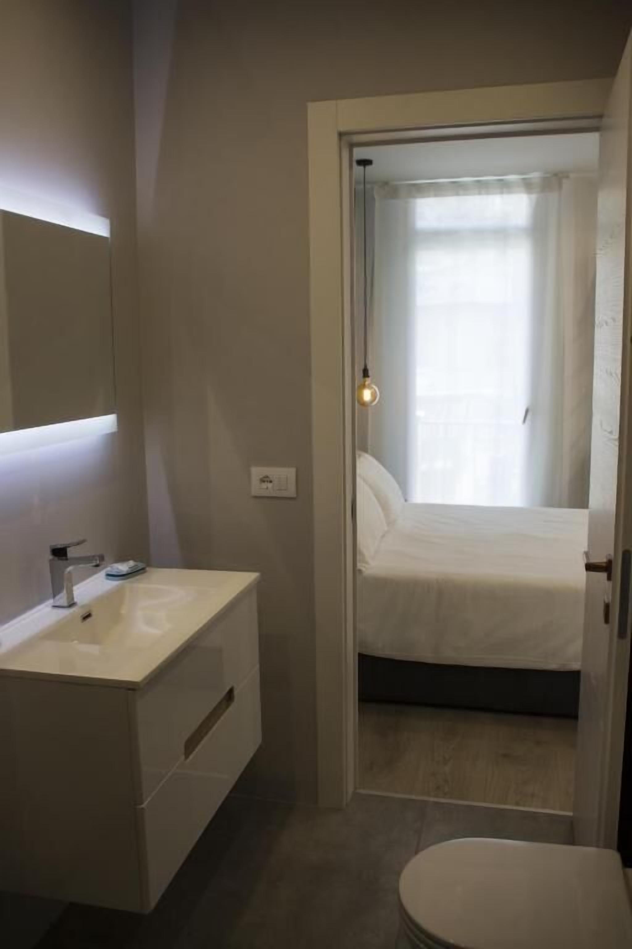 Un baño moderno con tocador blanco, inodoro y una cama visible a través de una puerta abierta.