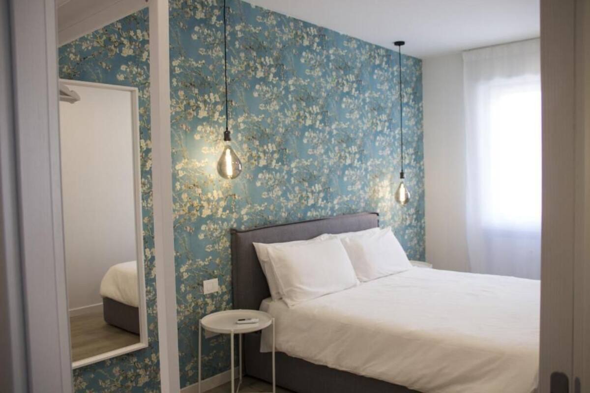 A bedroom with a bed, a bedside table, a mirror, and a wall with floral wallpaper.