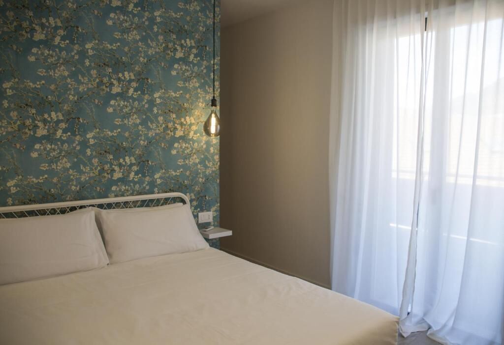 A bedroom with a bed, a nightstand, a wall with floral wallpaper, and a window with sheer curtains.