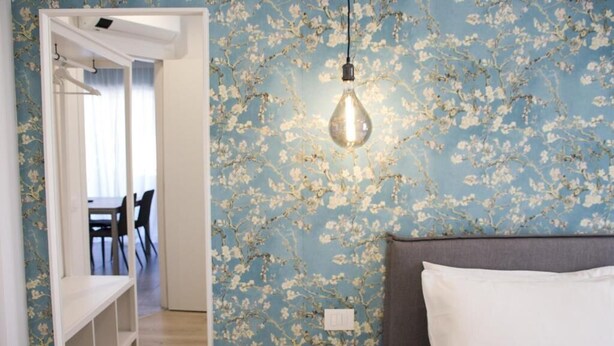 Un dormitorio con papel tapiz floral, una mesita de noche, un almohadón blanco y una lámpara colgante.