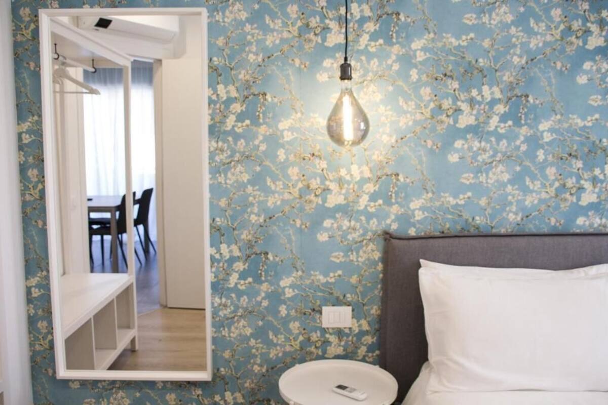 A bedroom with a floral wallpaper, a bedside table, a white pillow, and a hanging light fixture.