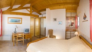 A bedroom with a wooden ceiling, a bed, a desk with a chair, a television, and a bathroom with a mirror and sink. - Maison 60 Mètres mer et Plage (Saint-Denis-d'Oléron)