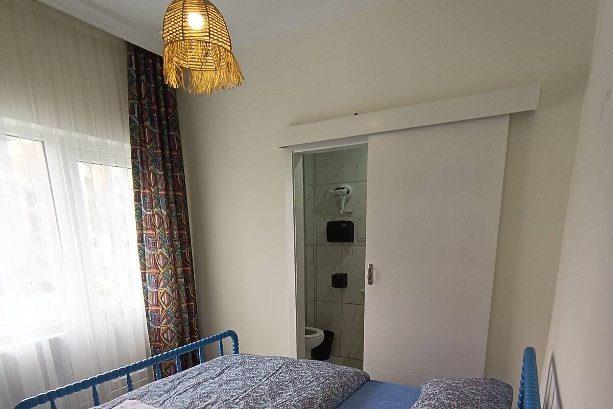Un dormitorio con cama azul, ropa de cama estampada, lámpara colgante, zona de ducha y ventana con cortinas.
