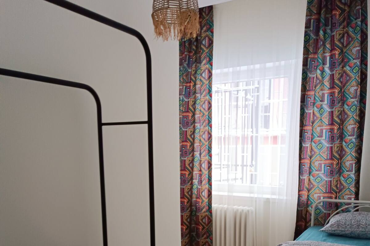 Un dormitorio con una cama litera, una mesita de noche con una cesta, una lámpara colgante, una ventana con cortinas estampadas y un radiador.