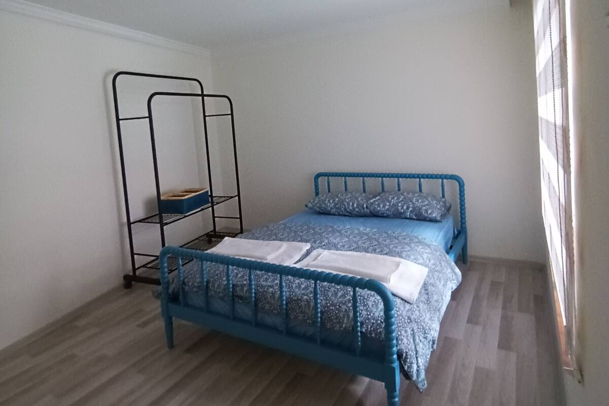 Una habitación con una cama de estructura metálica azul, una estantería de metal y suelo de madera.