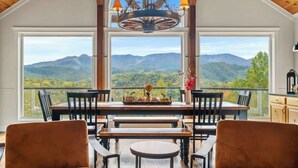 A dining area with a table and chairs, a chandelier, and a view of mountains through large windows. - Luxury Family Cabin | Hot Tub, Game Room & Views (Gatlinburg)