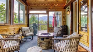 A cozy outdoor seating area with wicker furniture, a stone fire pit, and large windows offering mountain views. - Luxury Family Cabin | Hot Tub, Game Room & Views (Gatlinburg)