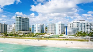 A beachfront hotel with multiple high-rise buildings, a sandy beach, and clear blue skies. - 2bd Beachfront Master Ste, Bay View w/balcony, Pool, nearby dining, beach access (Miami Beach)