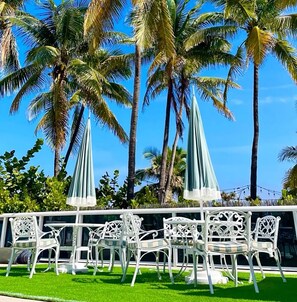A patio with white metal chairs and tables, surrounded by palm trees and a clear blue sky.