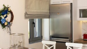 A modern kitchen with white cabinetry, a stainless steel refrigerator, and a wooden dining table with white chairs. - Elsa Villa by Elrin Villas Mani (West Mani)