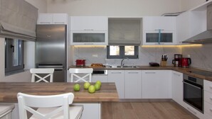 A modern kitchen with white cabinets, a stainless steel refrigerator, a wooden countertop, and a window with blinds. - Elsa Villa by Elrin Villas Mani (West Mani)