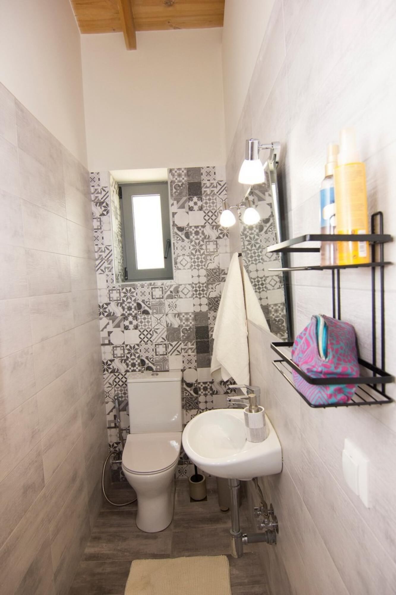 Un baño pequeño con inodoro, lavamanos y espejo con iluminación. Hay artículos de aseo en un estante y un salpicadero de azulejos estampados.