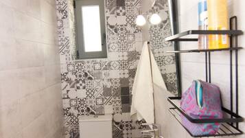 Un baño pequeño con inodoro, lavamanos y espejo con iluminación. Hay artículos de aseo en un estante y un salpicadero de azulejos estampados.