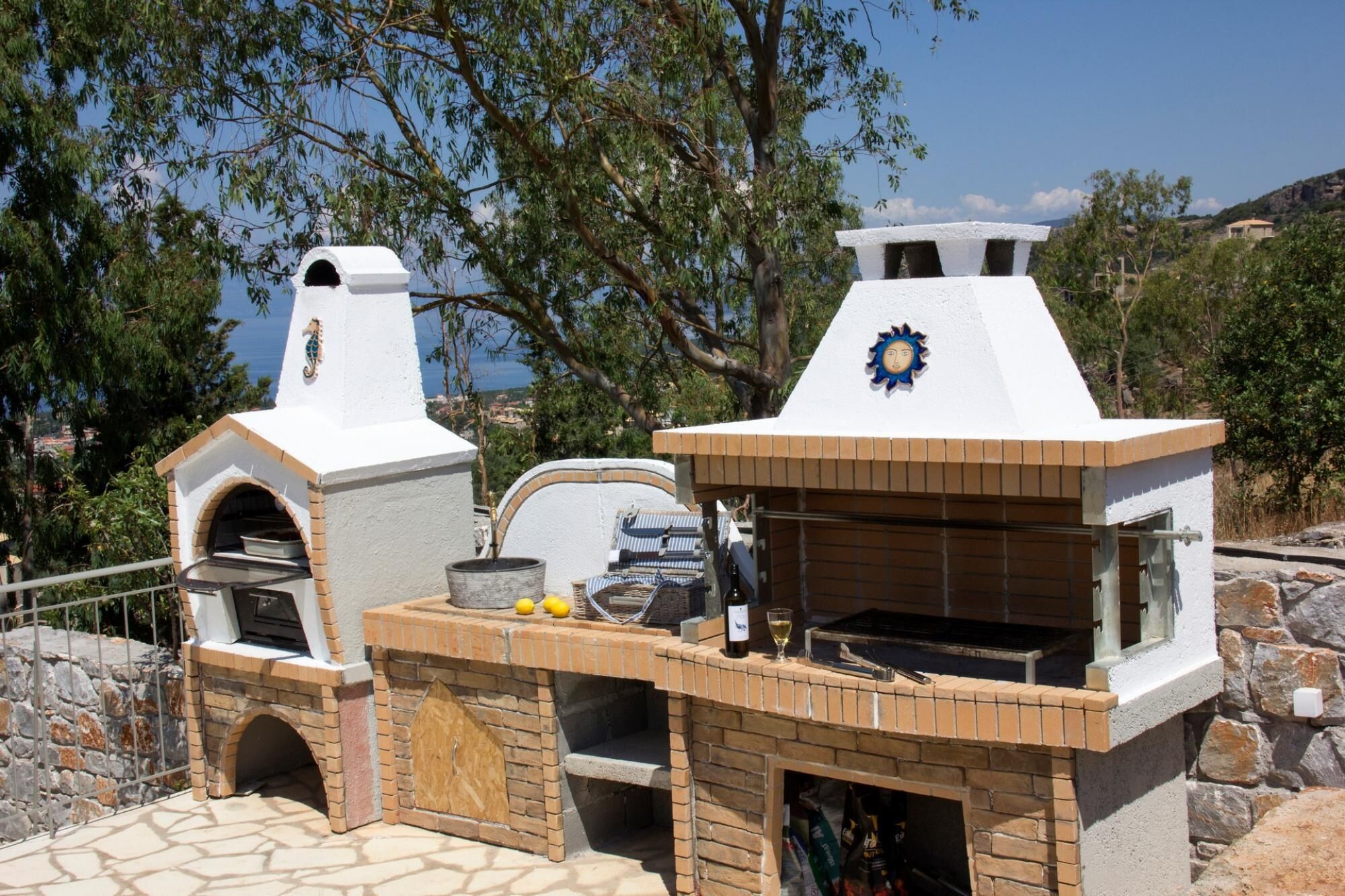 A stone patio with two brick ovens, a grill, and a small table with lemons and a bottle.