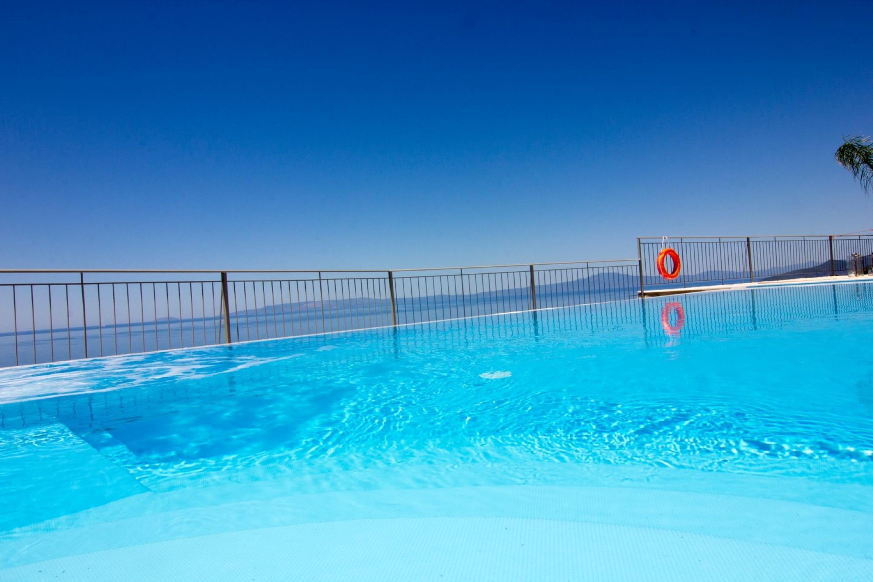 A clear blue swimming pool with a metal fence and a lifebuoy, surrounded by a clear sky.