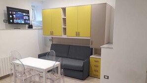 Una cucina moderna con mobili gialli e grigi, un divano grigio, un tavolo da pranzo bianco con sedie trasparenti e un televisore montato a parete.