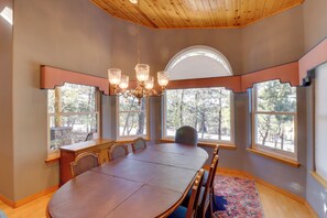 A dining area with a large wooden table, chairs, a chandelier, and large windows with views of trees. - Patio & Forest View: Peaceful Show Low Home (Show Low)