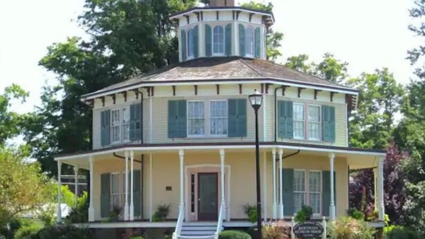 Une maison à deux étages dotée d’une véranda qui fait le tour de la maison, d’une tourelle et d’une entrée principale avec des marches.